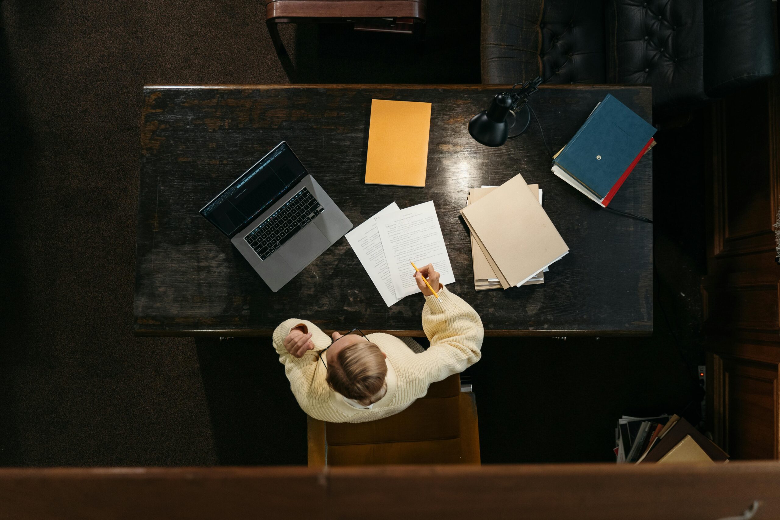 Person in yellow sweater working at a wooden desk with documents, folders, and a laptop.
